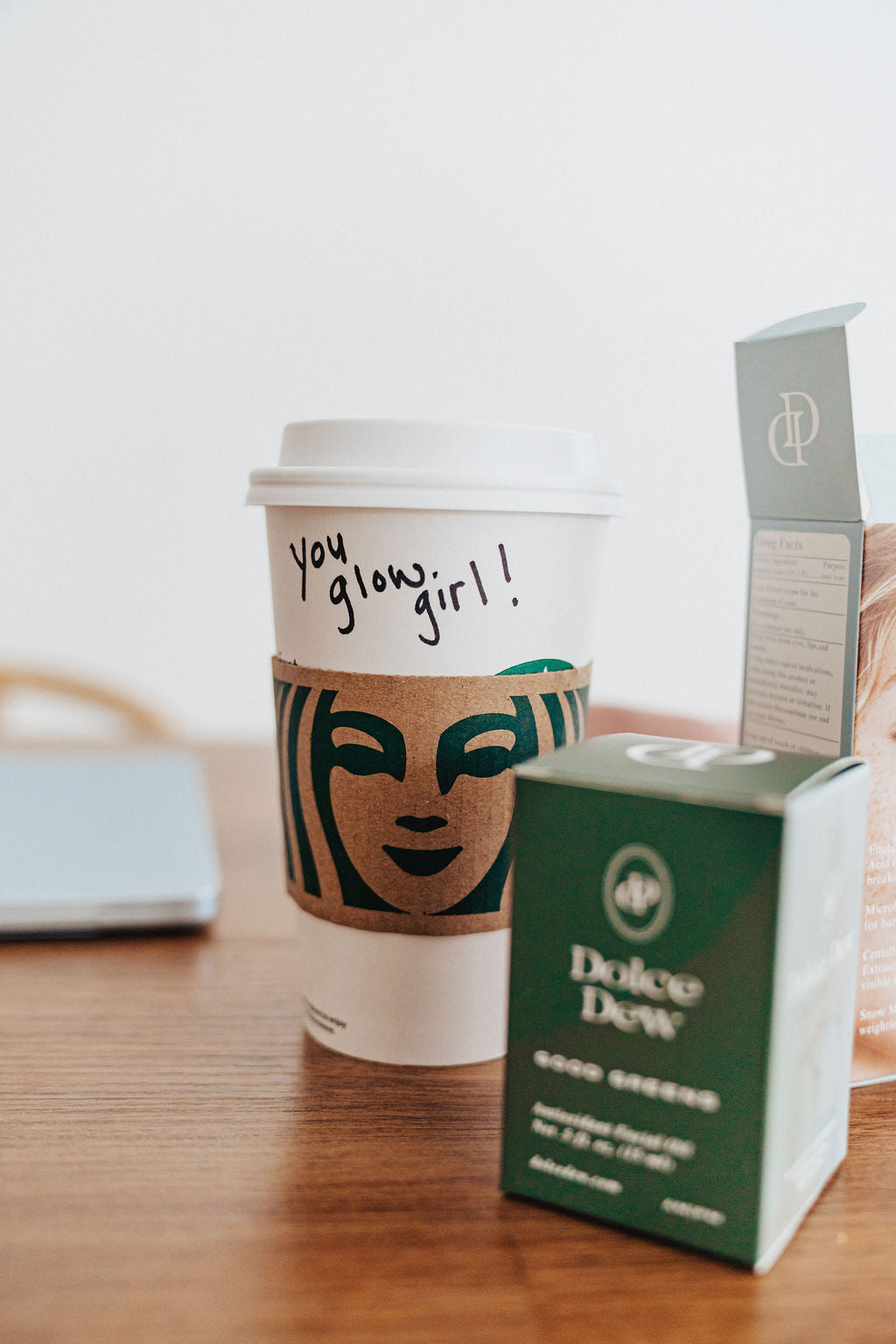 A white Starbucks paper coffee cup with a brown sleeve and handwritten text sits on a wooden table next to a green Dolce Dew good Greens  face oil 